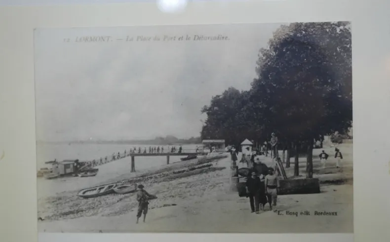 Photographie historique en noir et blanc du port et de l'esplanade de Lormont, avec des promeneurs, des bateaux amarrés et des arbres en arrière-plan, début XXe siècle.