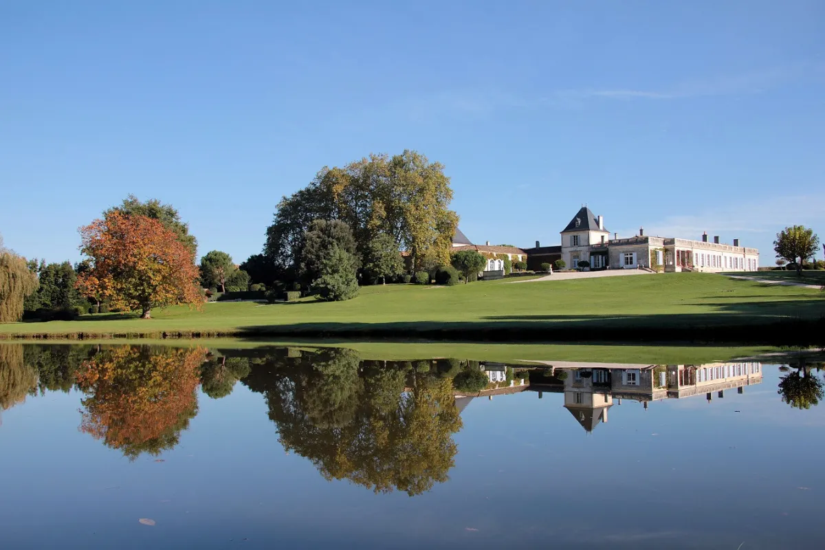 Vue du Château Paveil de Luze à Bordeaux avec sa façade blanche classique se reflétant dans l'étang, entouré d'arbres aux feuillages d'automne et d'une vaste pelouse verte sous un ciel bleu.