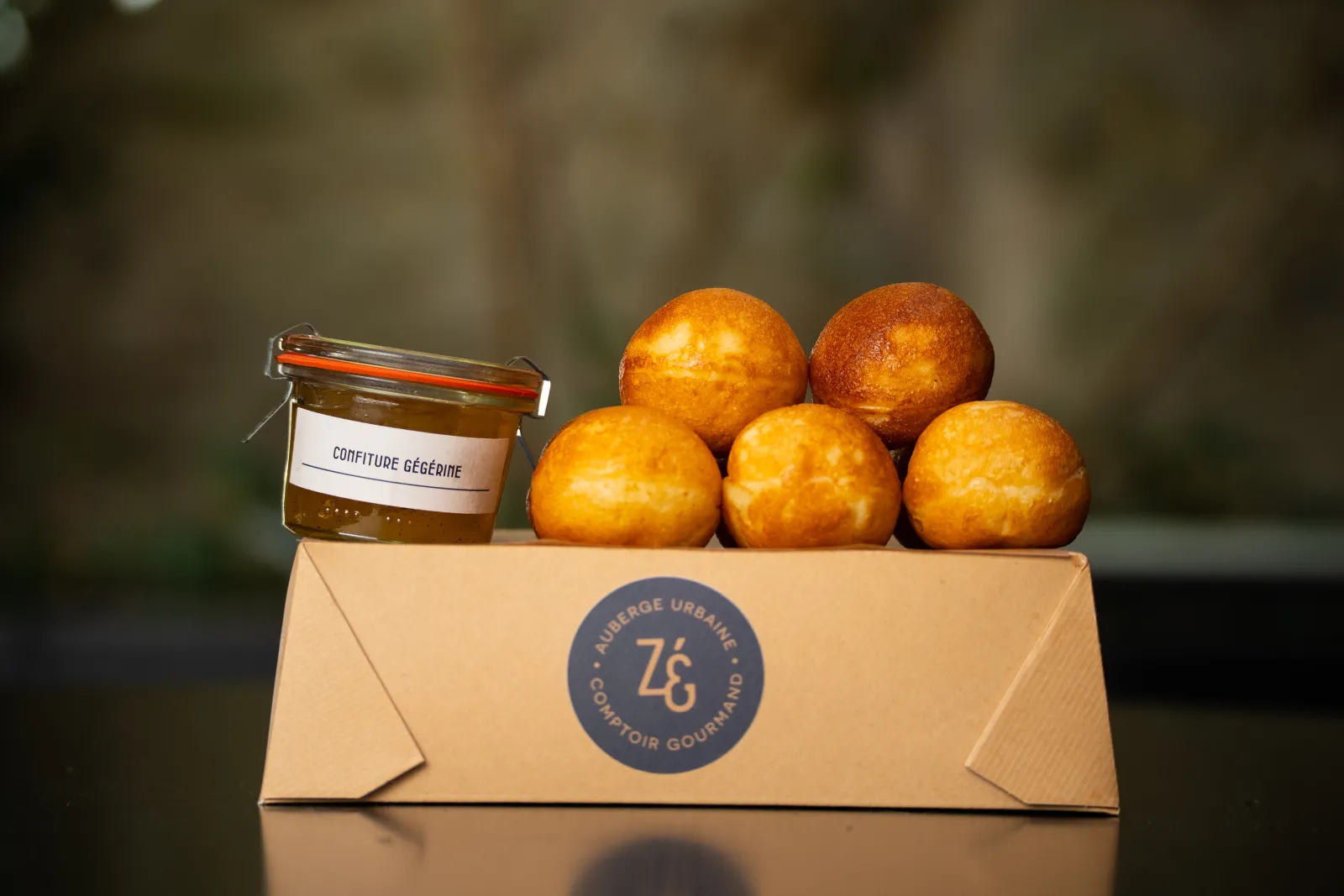 Boîte de présentation du Zeph' avec logo, contenant cinq pastels dorés et un pot de confiture Gégedin, sur fond flou.