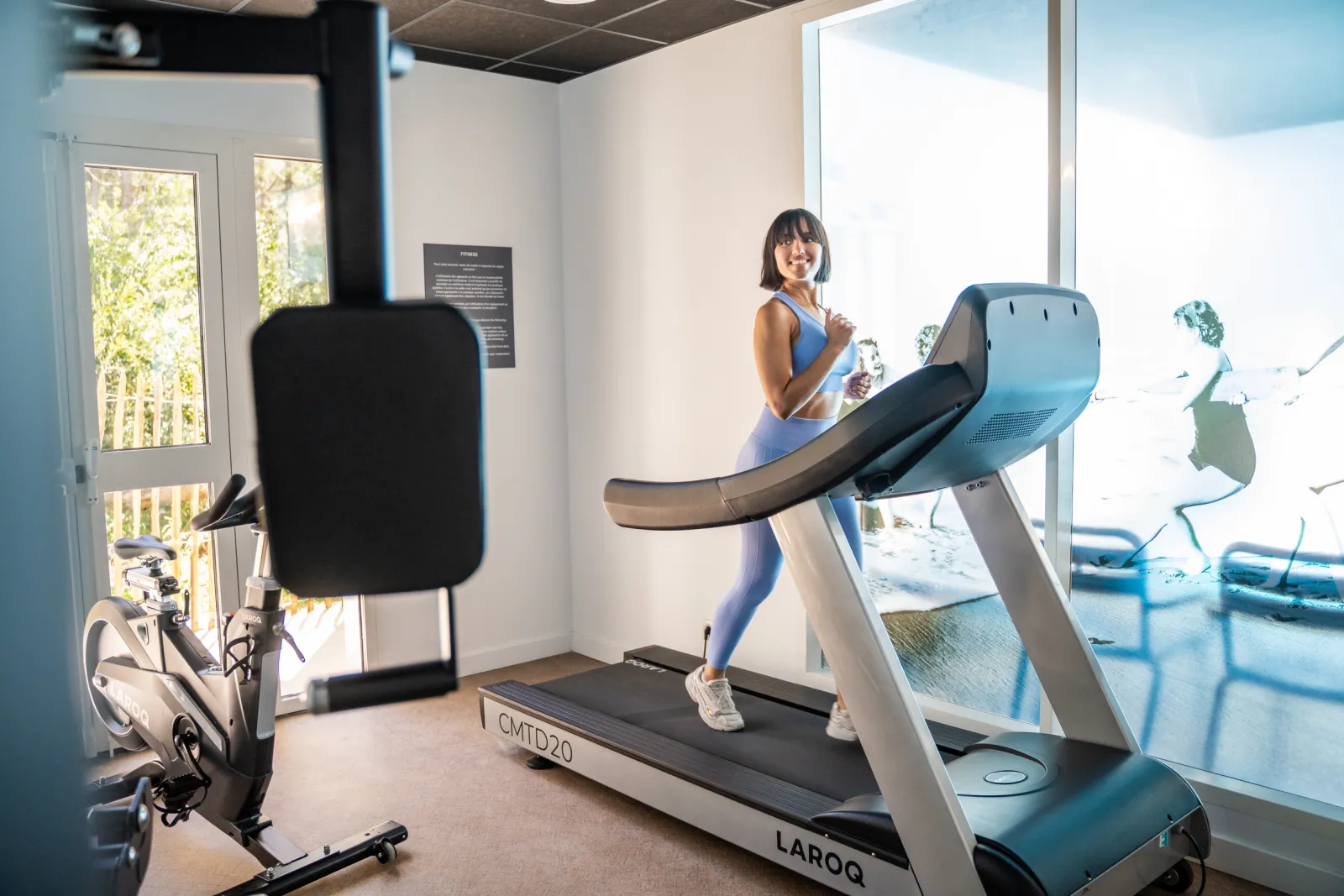 Femme en tenue de sport courant sur un tapis roulant LAROC dans une salle de fitness moderne avec vue sur Bordeaux, équipée d'un vélo d'appartement en arrière-plan