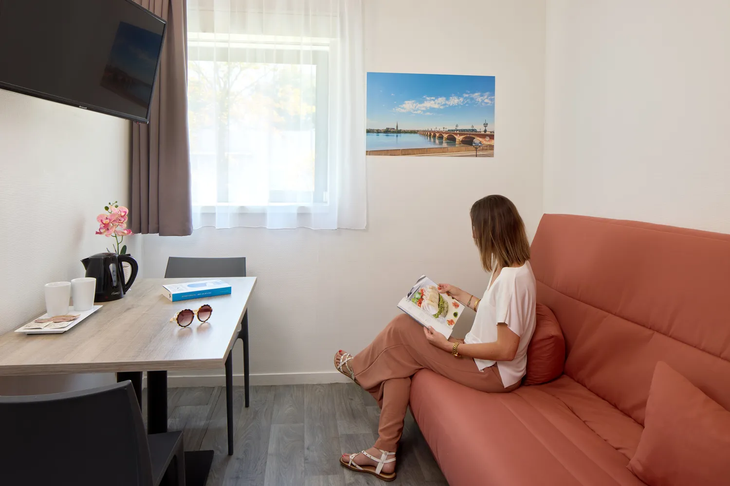 Une femme assise sur un canapé terracotta dans une chambre moderne, tenant une magazine de voyage, avec un bureau, une fenêtre lumineuse et une photo de Bordeaux au mur.