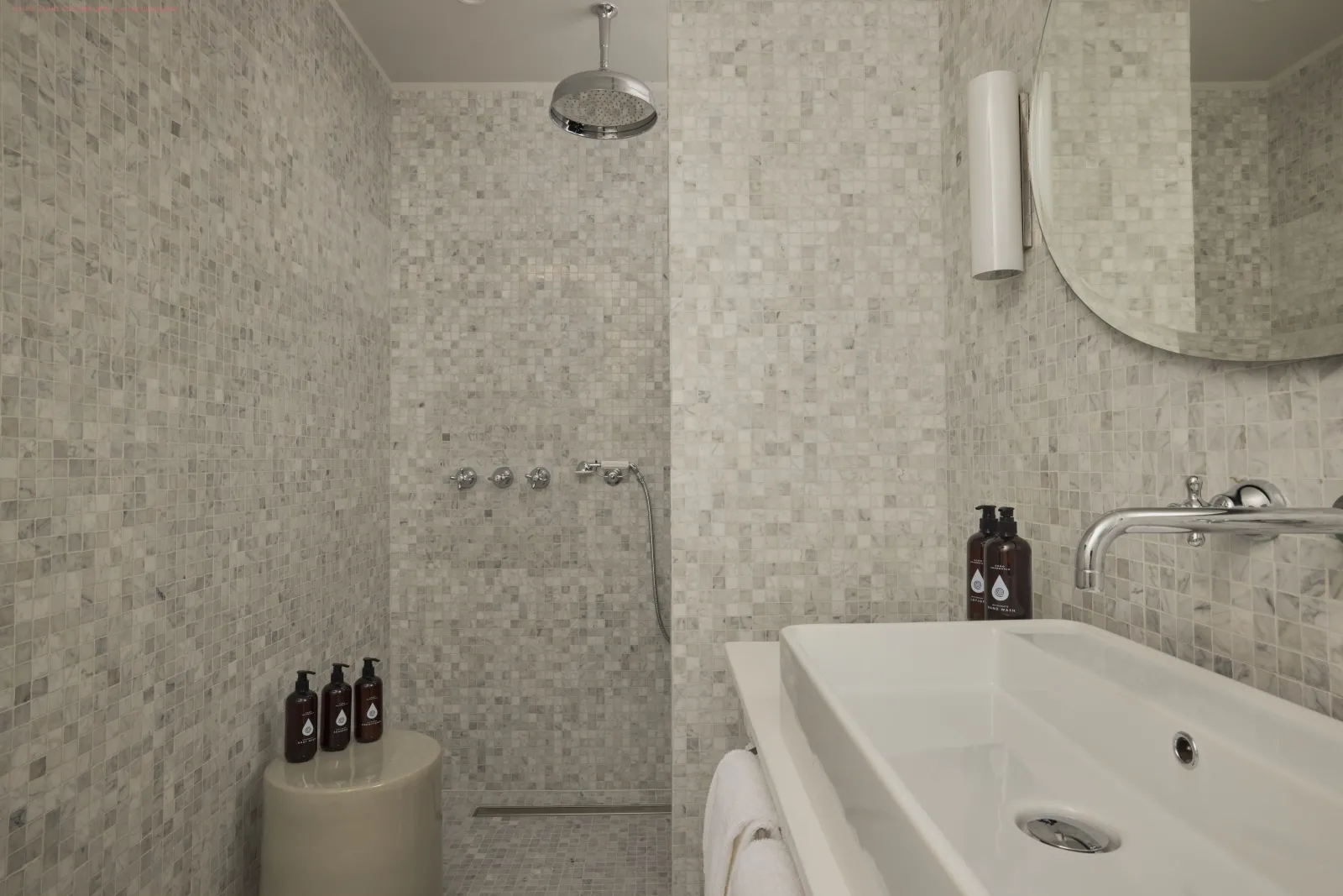 Salle de bain moderne avec douche à l'italienne, carrelage mosaïque beige, baignoire blanche, miroir rond et accessoires de luxe dans une suite du Como Cordeillan-Bages à Bordeaux