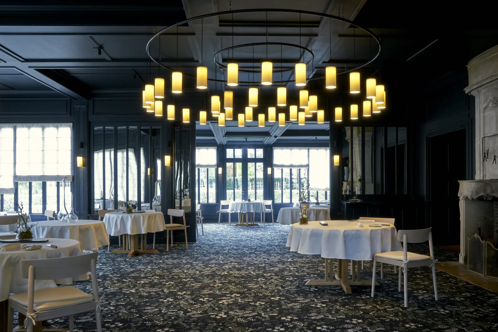Salle de restaurant élégante à la Grand'Vigne de Bordeaux avec tables rondes dressées, lustre moderne en cylindres lumineux, grandes baies vitrées et décoration contemporaine