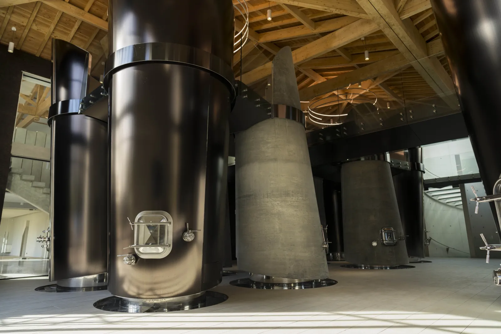 Grandes cuves de fermentation métalliques cylindriques dans une cave moderne du Château Montlabert à Bordeaux, avec poutres en bois apparentes au plafond.