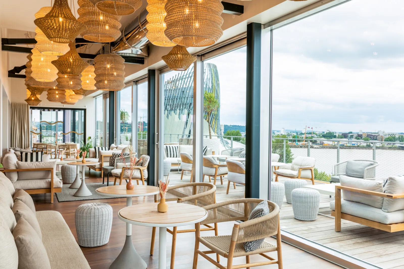 Intérieur moderne d'un restaurant ou lounge à Bordeaux avec vue panoramique sur la Garonne, luminaires dorés suspendus, mobilier contemporain en bois et tissu clair, terrasse extérieure aménagée.