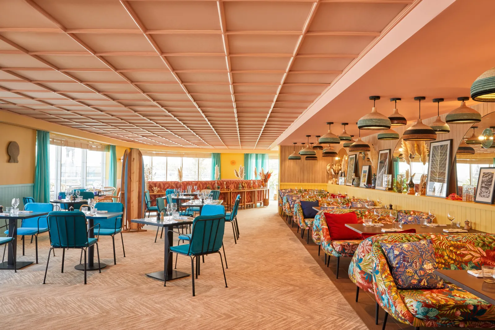 Restaurant moderne avec plafond géométrique en bois clair, chaises bleues, banquettes colorées aux motifs floraux, luminaires suspendus dorés et vues panoramiques sur Bordeaux