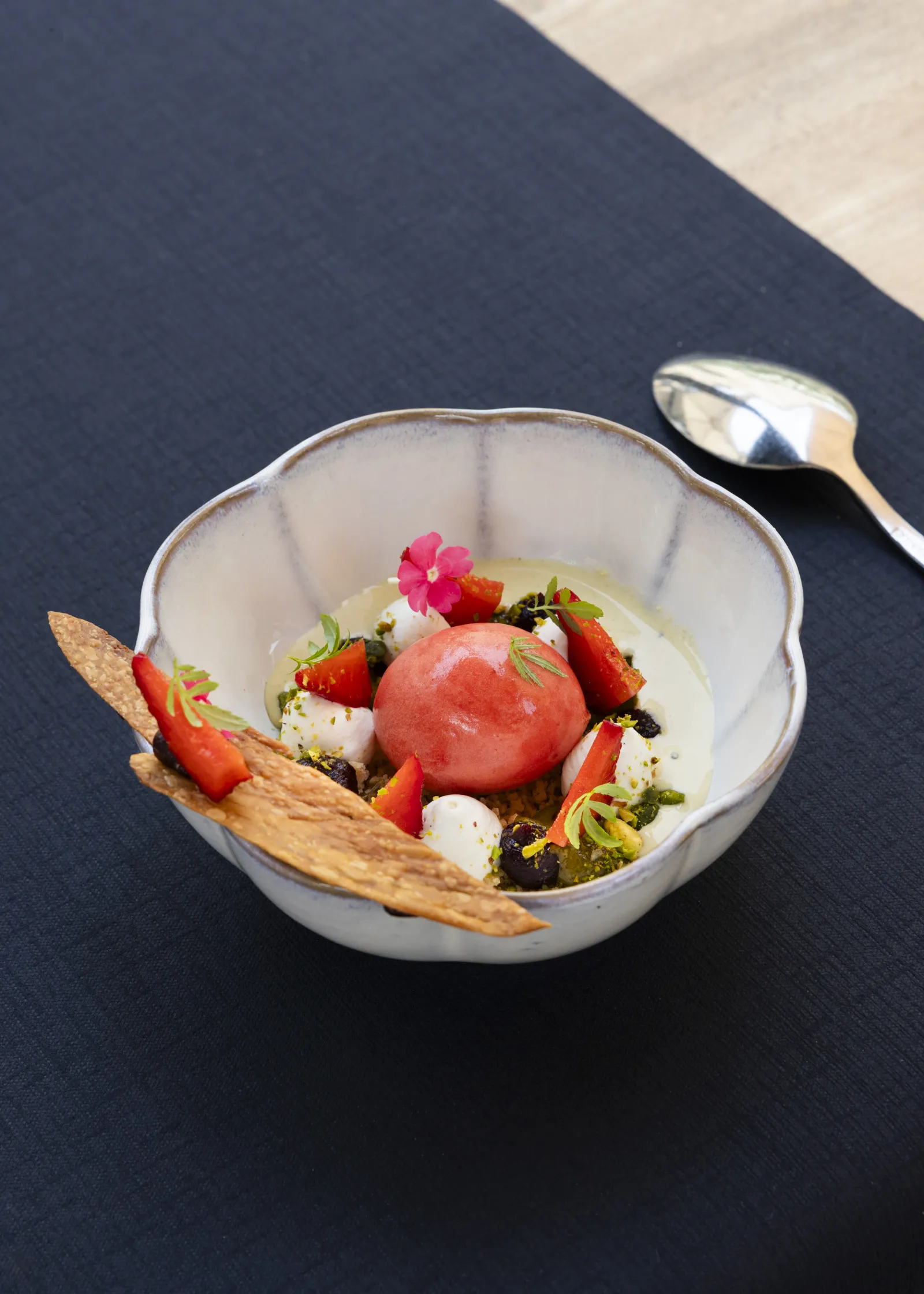 Assiette élégante de fine cuisine gastronomique présentant une boule de tomate rouge, des fraises, de la mozzarella blanche, des microgreens, une fleur comestible rose, et une tuile dorée, servie sur fond noir à la Terrasse de Lafaurie à Bordeaux.
