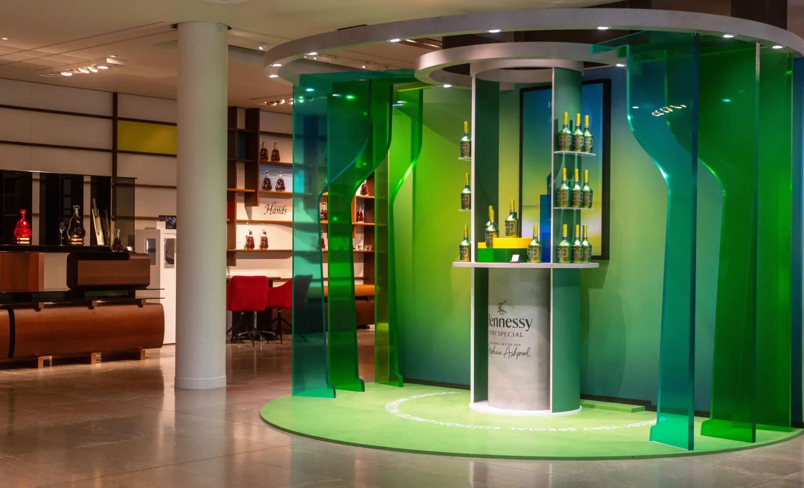Espace de visite moderne de la maison Hennessy à Bordeaux avec display circulaire en verre vert présentant des bouteilles de cognac, entouré de panneaux lumineux et de mobilier contemporain.