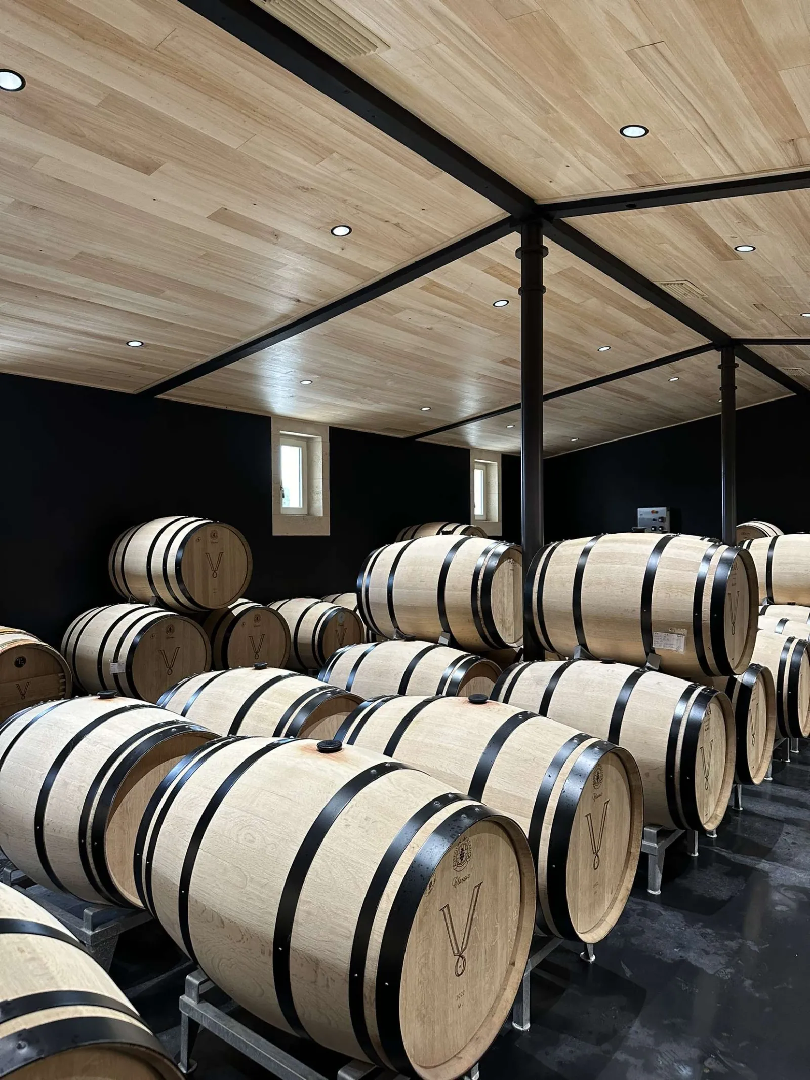 Cave de vieillissement avec rangées de barriques de vin en chêne alignées sous un plafond en bois clair et structure métallique noire, Château Petit Val à Bordeaux
