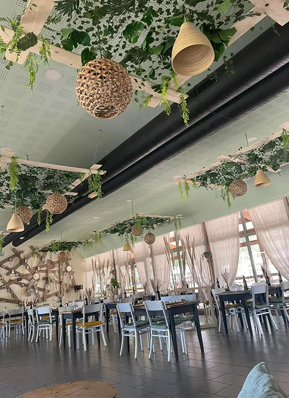 Intérieur du Bistrot du Lac à Bordeaux avec salle à manger spacieuse, chaises noires et blanches, grandes fenêtres avec rideaux blancs, décoration végétale suspendue et luminaires en osier naturel.