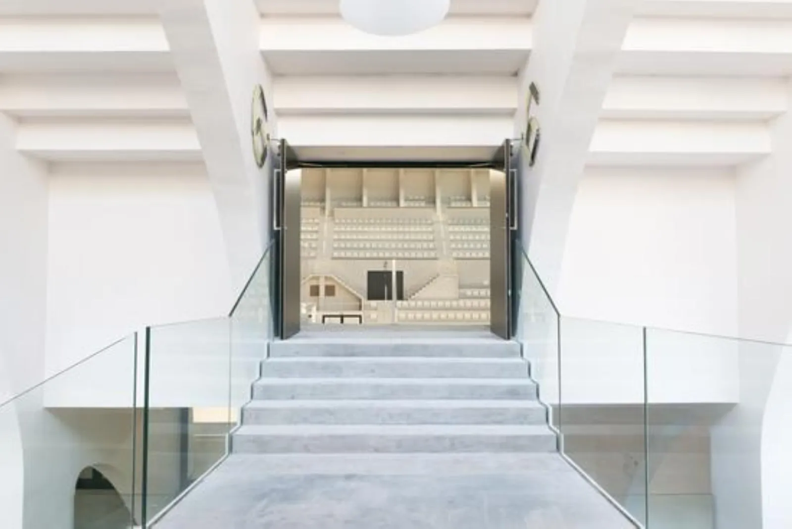 Vue symétrique d'un escalier moderne montant vers les portes d'entrée du Palais des Sports de Bordeaux, avec rampes en verre, plafond blanc géométrique et architecture épurée
