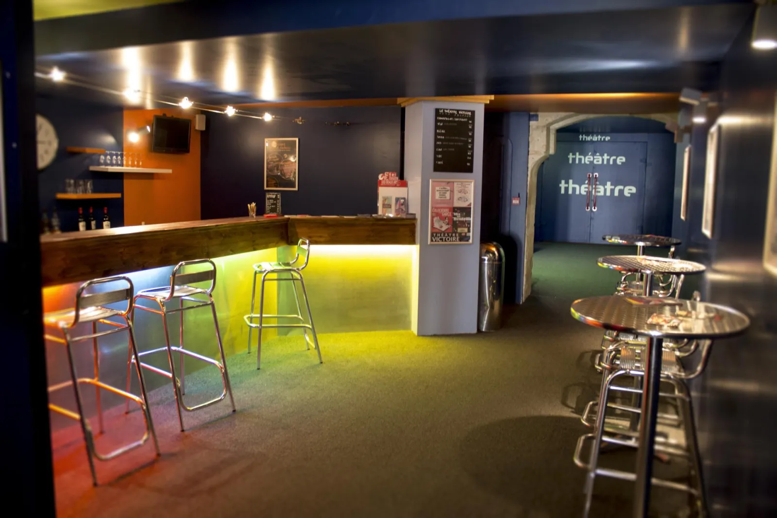 Intérieur du Théâtre Victoire de Bordeaux avec un bar luminescent jaune-vert, des tabourets métalliques, des tables hautes, et l'entrée de la salle de théâtre visible en arrière-plan avec sa signalétique bleue.