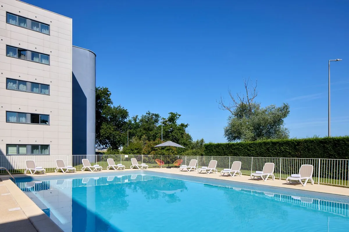 Piscine extérieure de l'hôtel Zenitude Bordeaux Aéroport avec transats blancs, bâtiment moderne en arrière-plan et ciel bleu dégagé.