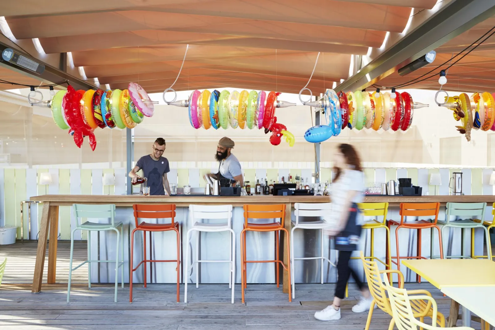 Bar du Mama Shelter à Bordeaux avec un comptoir en bois, des chaises colorées et des bracelets fluorescents suspendus au plafond en bois. Deux barmen servent les clients dans un espace lumineux et moderne.