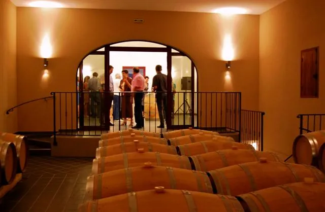 Cave de dégustation du Château Pichon Bellevue à Bordeaux avec tonneaux de chêne alignés au premier plan et visiteurs observant depuis une plateforme surplombante dans une salle aux murs ocre et éclairage chaleureux.