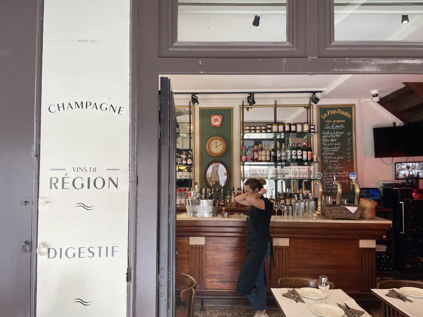 Intérieur d'une cave à vin bordelaise avec comptoir en bois, étagères de bouteilles, pancarte 'Champagne Vins de Région Digestif' à l'entrée, et une femme en noir derrière le bar.