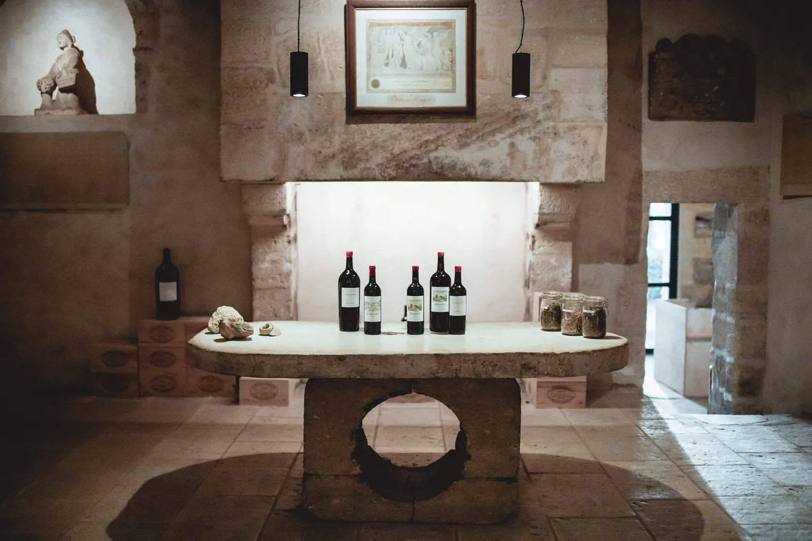 Intérieur du Château Mangot à Bordeaux : une table ronde en pierre avec des bouteilles de vin rouge et des bocaux, dans une cave aux murs de pierre patinés, éclairée par des lampes suspendues noires.