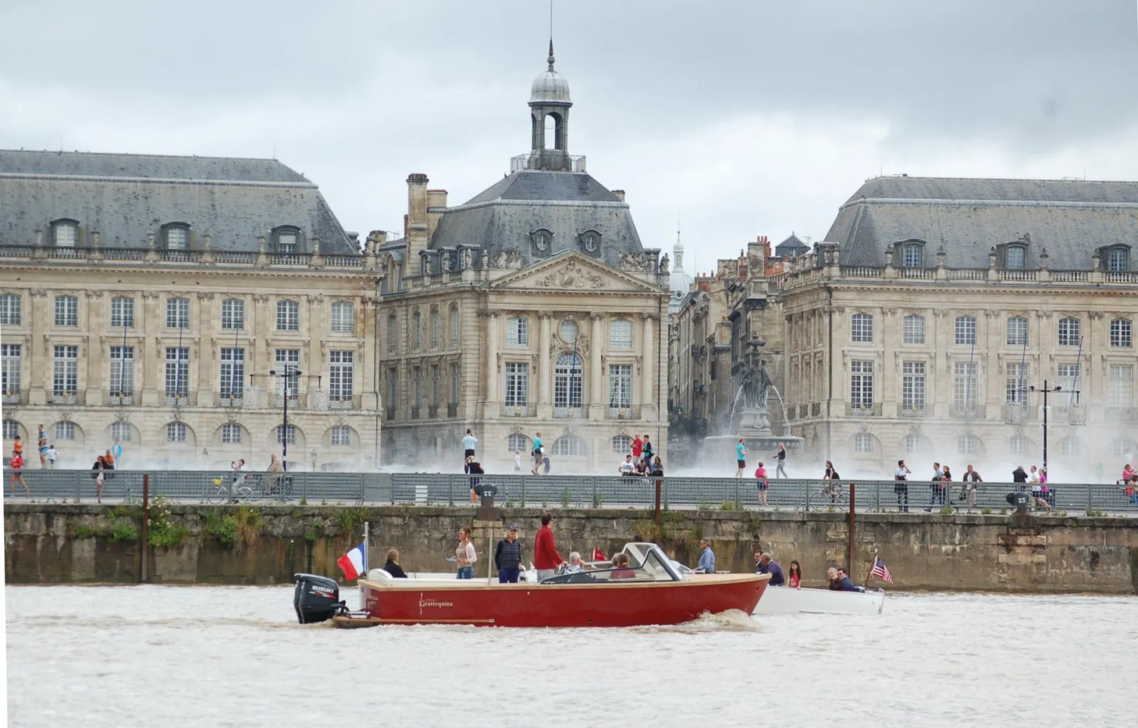 Le bateau taxi rouge Grattequina navigue sur la Garonne à Bordeaux, avec en arrière-plan la Place de la Bourse et ses façades classiques du XVIIIe siècle, sous un ciel nuageux.
