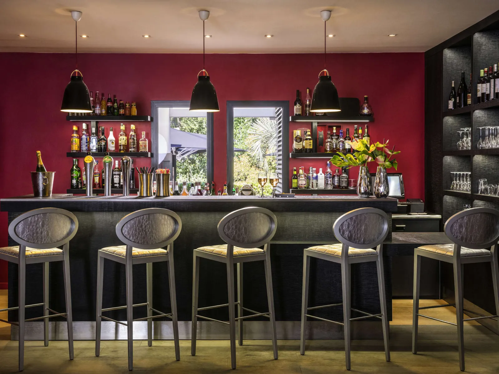 Un bar élégant avec un mur magenta, un comptoir noir avec cinq tabourets gris, trois lampes suspendues noires, des étagères de bouteilles d'alcool, et des baies vitrées donnant sur un paysage verdoyant.