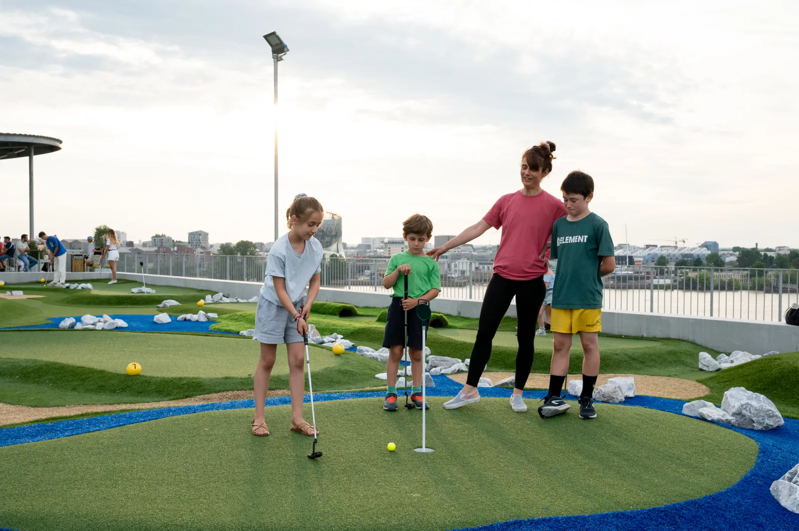 Une femme et trois enfants jouent au mini-golf sur un terrain synthétique avec vue sur Bordeaux en arrière-plan, à la UCPA Sport Station.