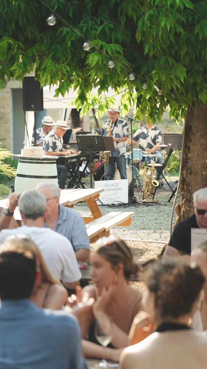 Soirée guinguette en plein air au Château Rol Valentin à Bordeaux, avec un groupe de jazz jouant sous un arbre feuillu, des spectateurs assis sur des bancs en bois écoutant le concert.