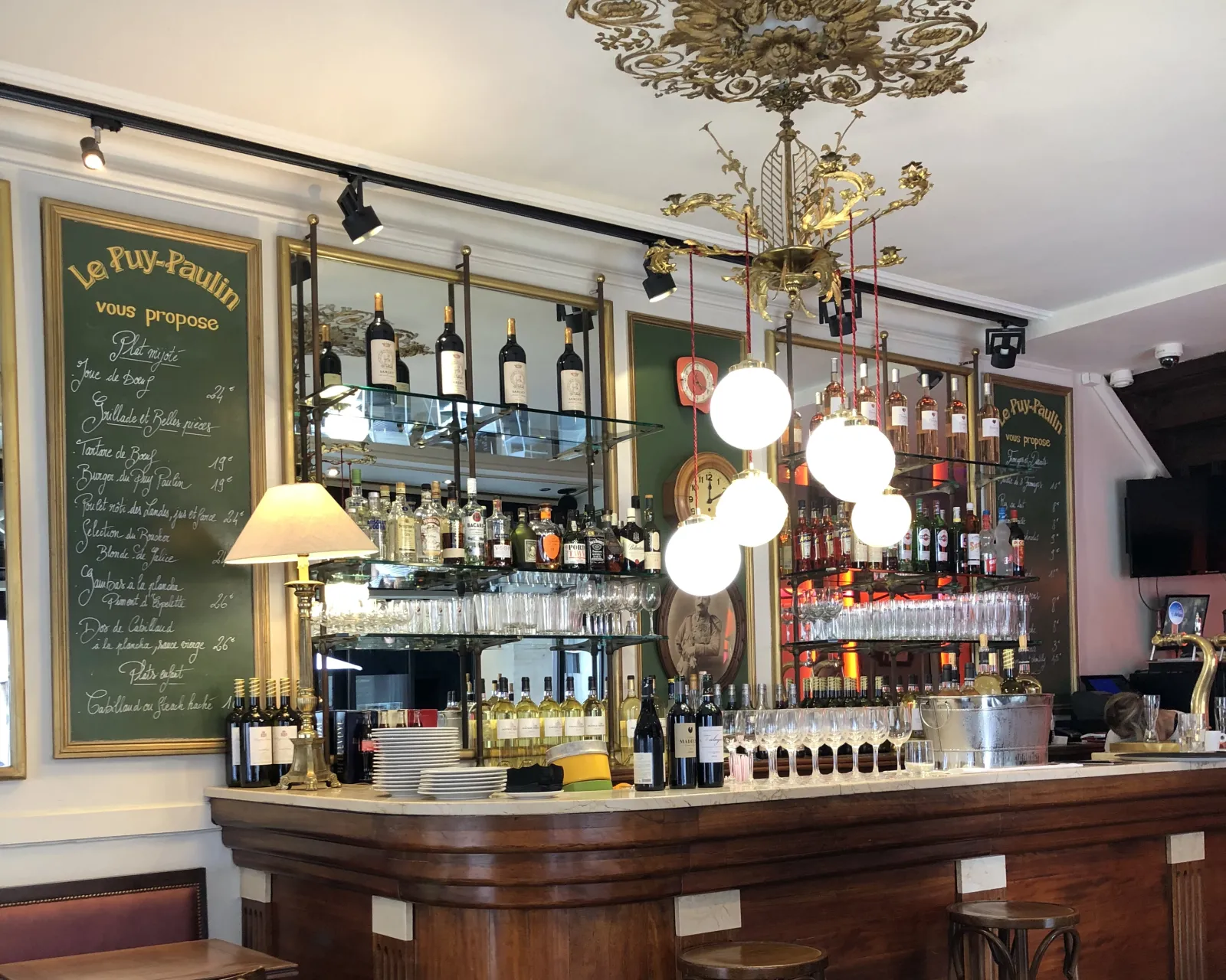 Le comptoir élégant du bar Le Puy-Paulin à Bordeaux, avec bouteilles de vin, verres, luminaires suspendus modernes et tableaux noirs affichant les propositions du jour.