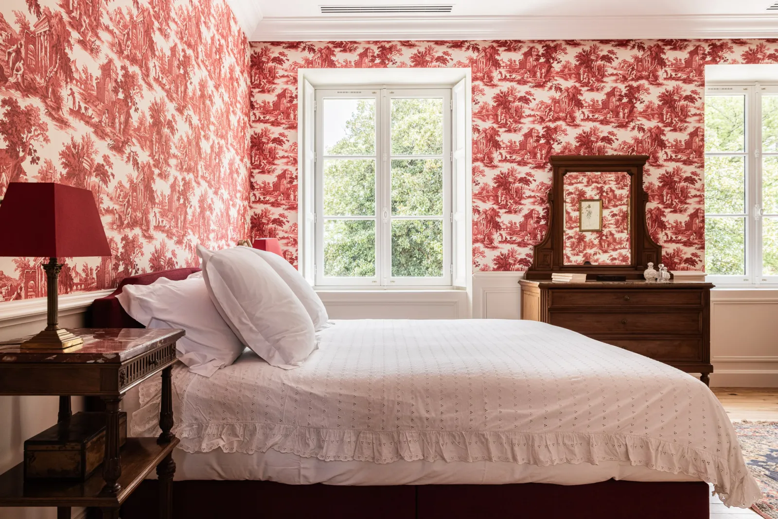Chambre élégante du Château Sénéjac à Bordeaux avec papiers peints rouge toile de Jouy, lit en bois massif, mobilier d'époque et grandes fenêtres donnant sur le parc.