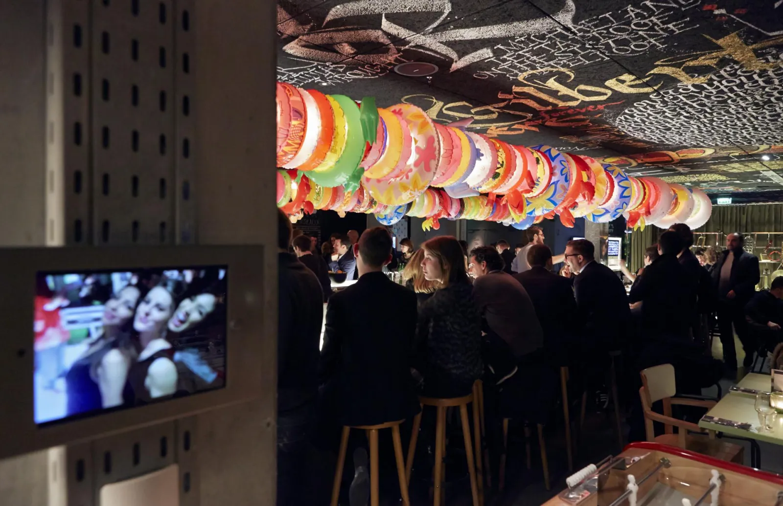 Intérieur du Mama Shelter Bar à Bordeaux avec des lanternes colorées suspendues au plafond, des clients en tenue noire se minglant dans une atmosphère festive, un écran TV visible à gauche et des graffitis décoratifs sur le plafond