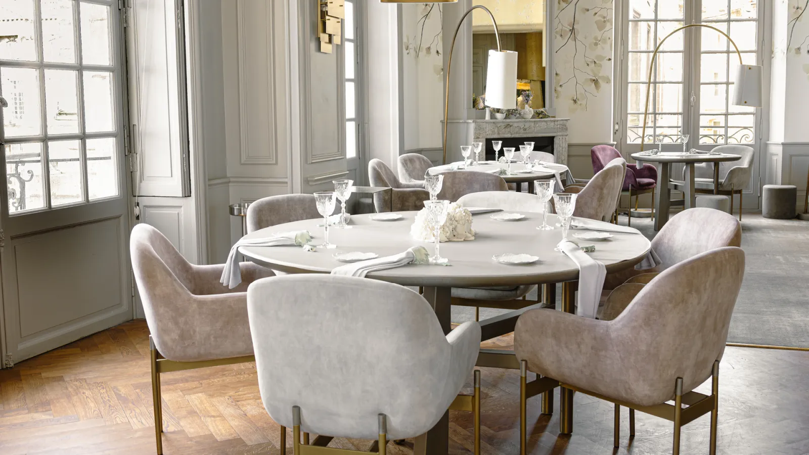 Salle de restaurant élégante avec tables rondes dressées, chaises beiges, grandes fenêtres donnant sur Bordeaux, miroir doré et décoration épurée.