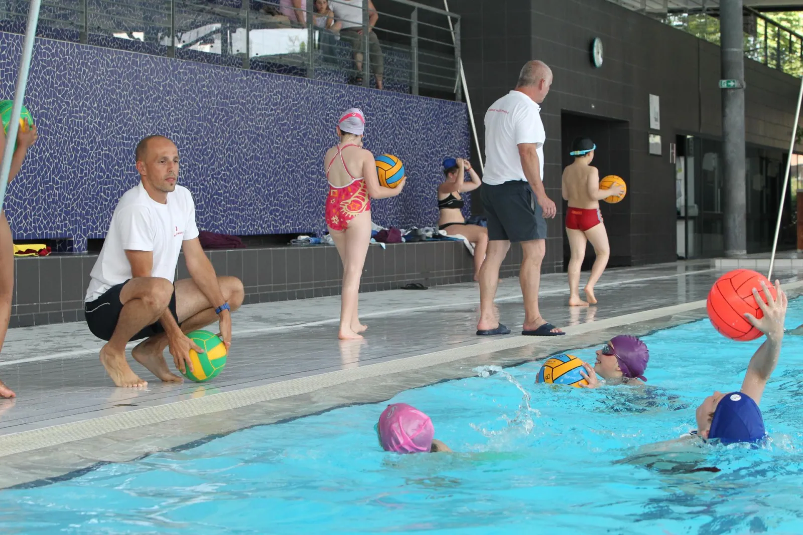 Cours de water-polo à la piscine municipale Tissot de Bordeaux avec un entraîneur et plusieurs enfants en maillot de bain pratiquant le sport dans l'eau et au bord du bassin.
