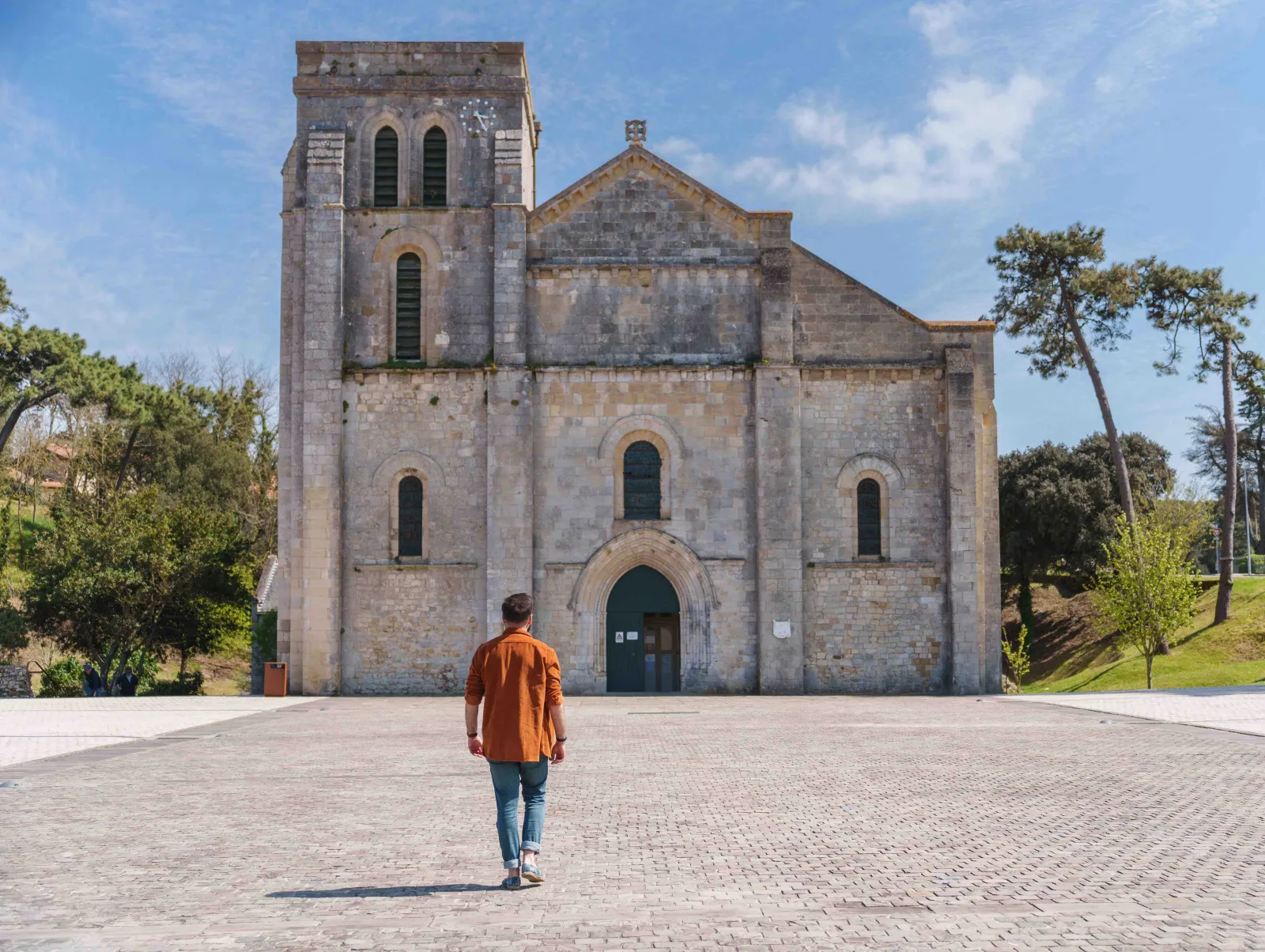 Un visiteur en chemise orange marche vers la Basilique Notre-Dame-de-la-fin-des-Terres à Bordeaux, édifice religieux en pierre blanche avec un clocher carré et une façade romane, entouré d'arbres sous un ciel bleu.