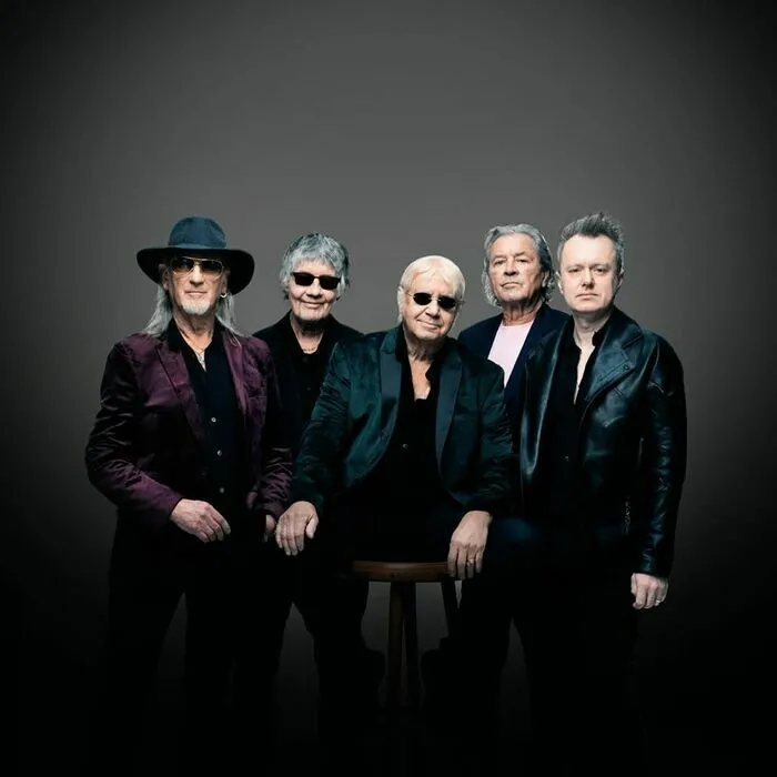 Portrait de groupe de cinq musiciens de Deep Purple en tenue de scène noire et accessoires, posant sur fond gris uni.