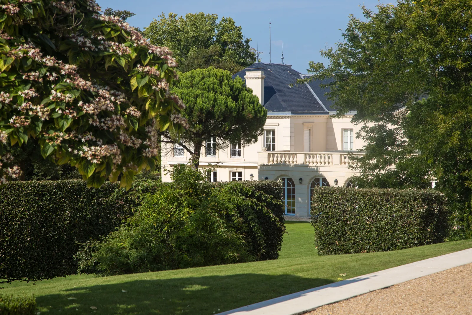 Vue du Château Malartic-Lagravière à Bordeaux, édifice blanc du XIXe siècle avec toit ardoisé, entouré de haies taillées et de jardins à la française, avec des fleurs roses en premier plan.