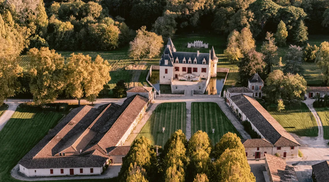 Vue aérienne du Château Olivier à Bordeaux avec ses vignobles en terrasses, son architecture médiévale avec tours et créneaux, entouré de forêts d'automne et de jardins à la française