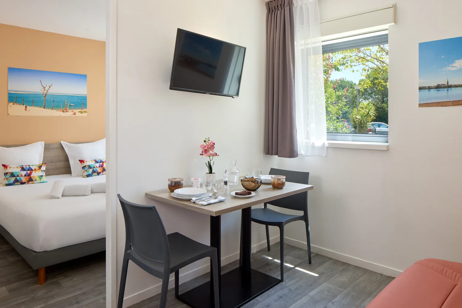 Chambre moderne d'hôtel à Bordeaux avec lit confortable, bureau, chaises noires, télévision murale, fenêtre avec vue sur la végétation, décoration côtière et mobilier contemporain.