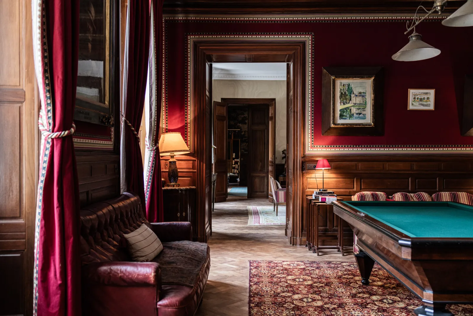 Salle de billard élégante du Château Sénéjac à Bordeaux avec murs bordeaux, boiseries en bois foncé, table de billard verte, fauteuil Chesterfield en cuir, rideaux rouges et portes ouvertes donnant sur des pièces adjacentes.