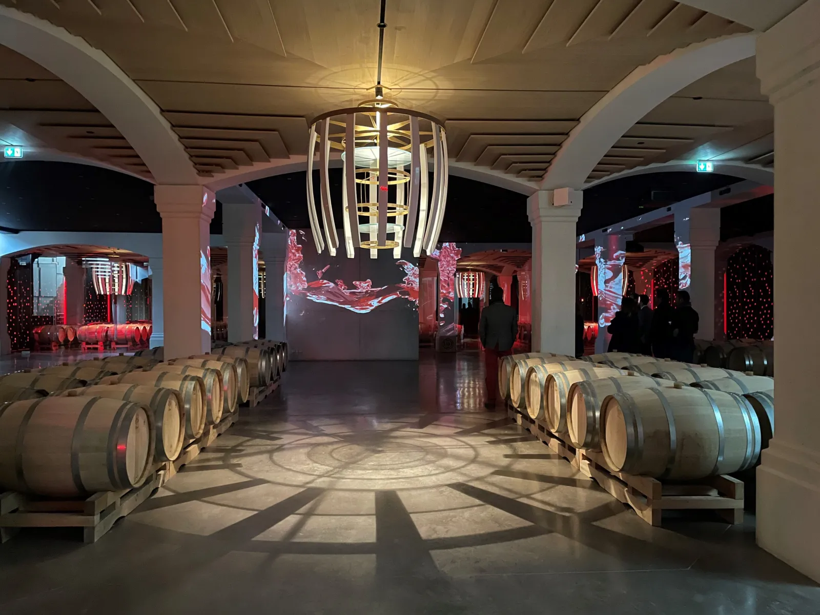 Cave de dégustation du Château Montlabert à Bordeaux avec tonneaux de vin alignés de part et d'autre d'une allée centrale, éclairage rouge ambiant et lustre moderne au plafond.