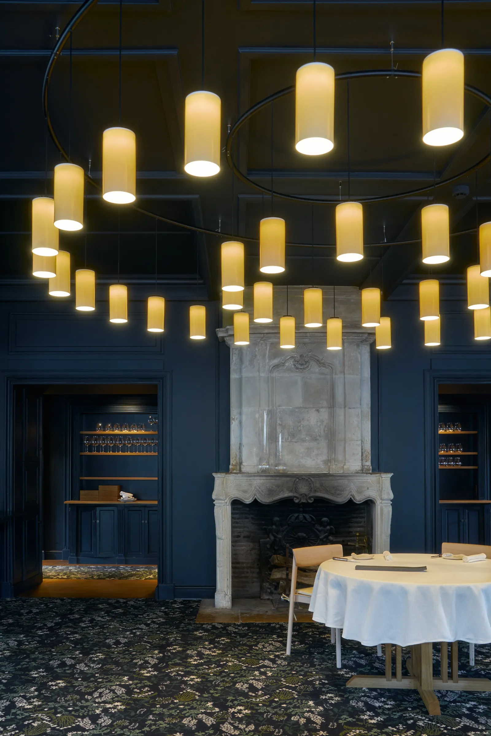 Intérieur élégant d'un restaurant avec plafond orné de luminaires cylindriques jaunes, murs bleu foncé, cheminée blanche, mobilier minimaliste et table ronde blanche au premier plan