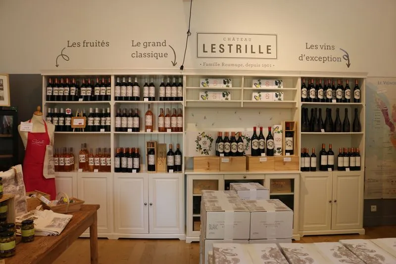 Vitrine de présentation du Château Lestrille à Bordeaux, avec des étagères organisées en trois sections : les fruits, le grand classique et les vins d'exception. Des bouteilles de vin sont exposées aux côtés de produits dérivés, avec des cartons blancs en