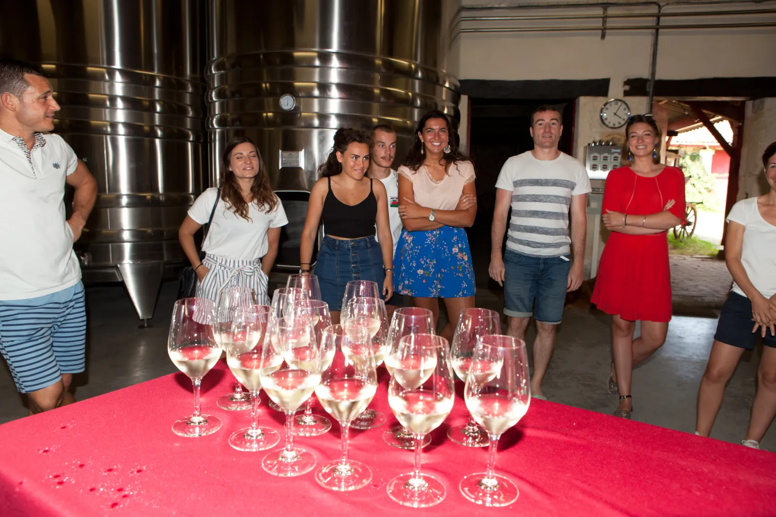 Groupe de visiteurs debout autour d'une table avec des verres de vin blanc devant une cuve de vinification en acier inoxydable dans une cave bordelaise.