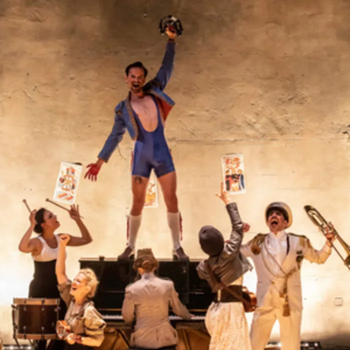 Scène de théâtre de l'opéra Carmen à Bordeaux : un danseur en costume bleu lève le poing au-dessus de sa tête, entouré de musiciens et de figurants jouant des instruments et tenant des affiches, sur fond de décor épuré.