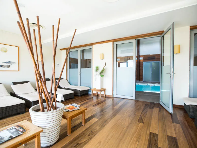 Espace spa luxueux avec chaises longues noires, décoration en bambou, baies vitrées donnant sur une piscine turquoise, sol en bois clair et mobilier en bois naturel aux Bains de Margaux à Bordeaux