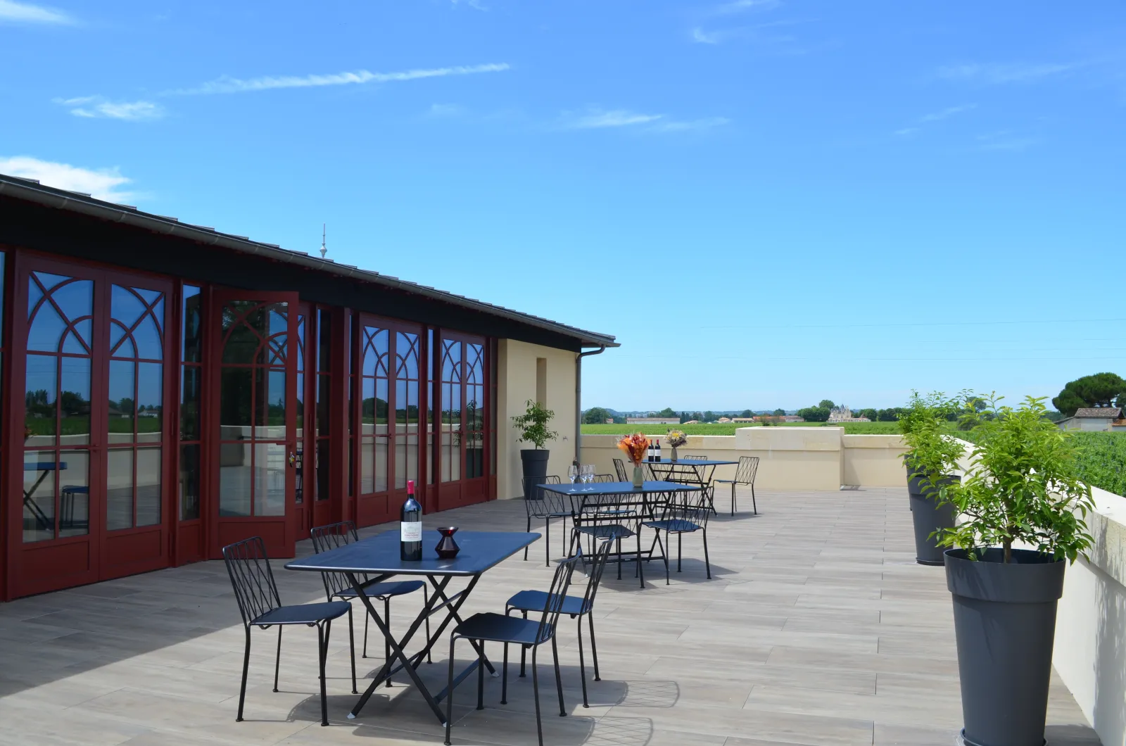 Terrasse ensoleillée du Château Yon-Figeac à Bordeaux avec tables et chaises, bâtiment aux portes rouges et vue sur les vignobles