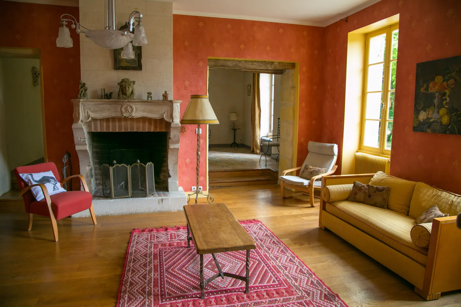 Salon élégant du Château Valoux à Bordeaux avec murs terracotta, cheminée blanche, mobilier vintage jaune et rouge, parquet bois, tapis rose et portes jaunes donnant sur le jardin