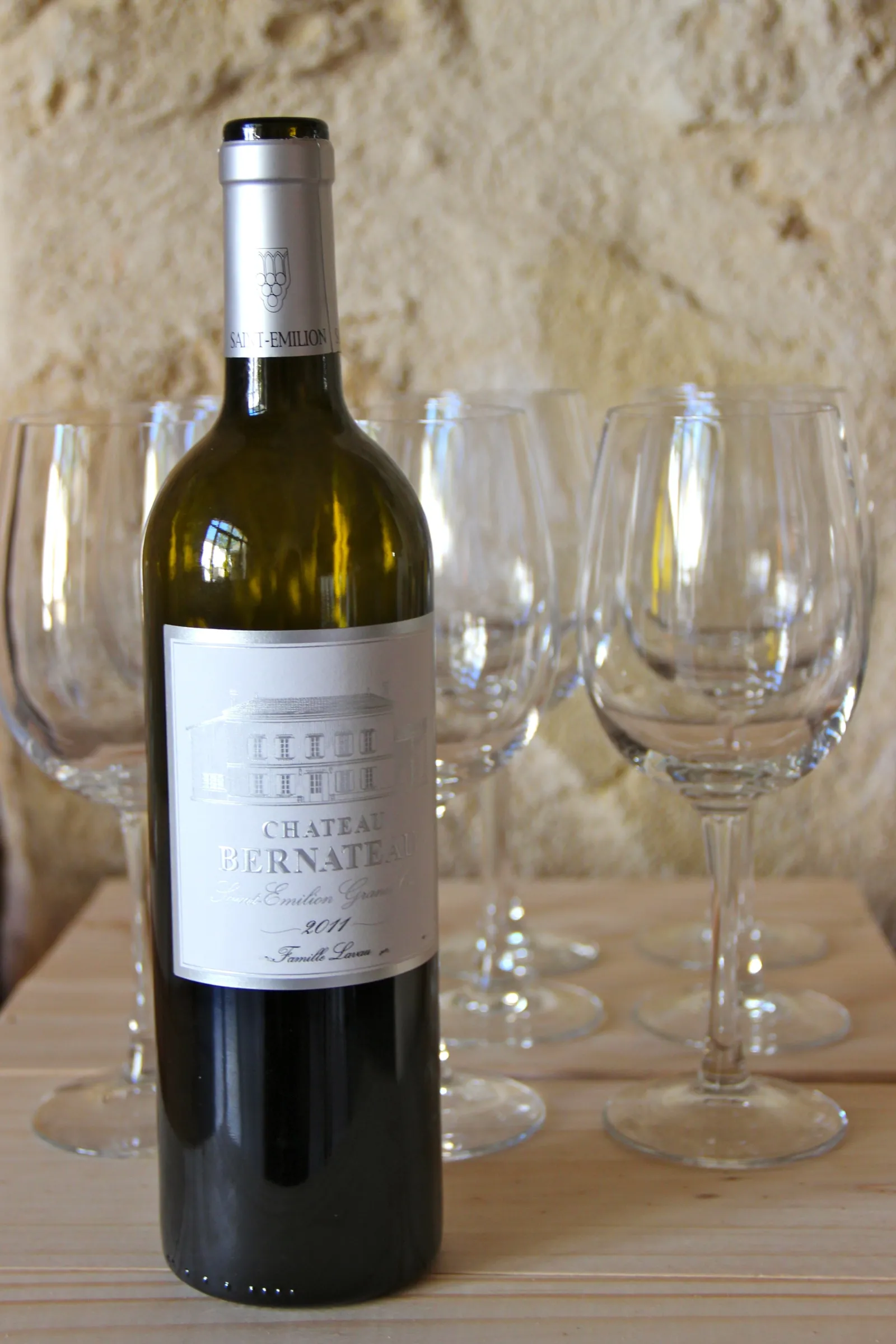 Bouteille de vin rouge Château Bernateau de Bordeaux entourée de verres à vin vides sur une table en bois, devant un mur en pierre.