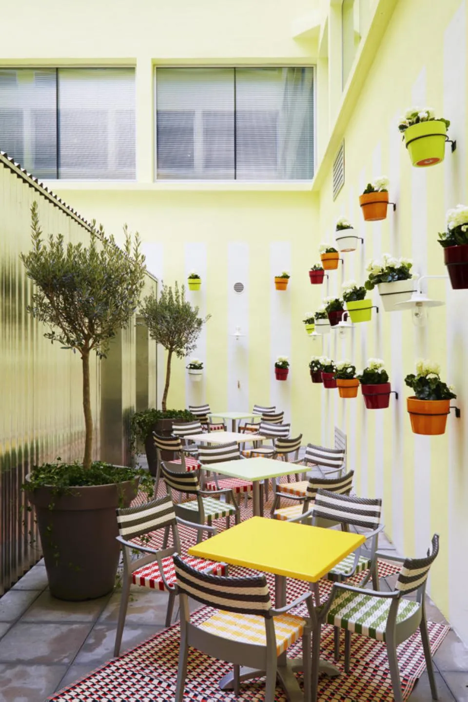 Terrasse couverte du Mama Shelter Restaurant à Bordeaux avec murs jaune vif, chaises rayées bleu et blanc, tables colorées, pots de fleurs suspendus multicolores et plantes en pots.