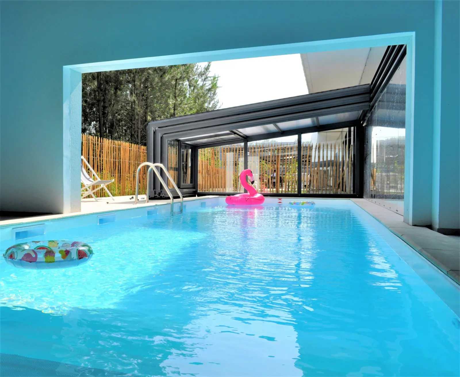 Piscine couverte intérieure avec eau turquoise, flamant rose gonflable, accès à la terrasse extérieure avec bois et forêt en arrière-plan, Holiday Inn Bordeaux Mérignac