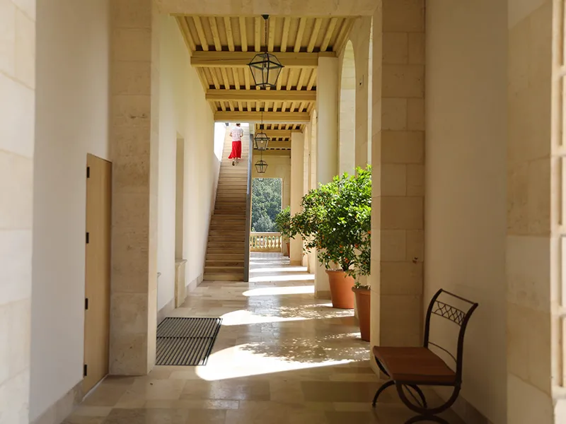 Galerie couverte du Château Marquis d'Alesme Becker à Bordeaux, avec colonnes blanches, plafond à claire-voie en bois, plantes en pots, et escalier visible en arrière-plan