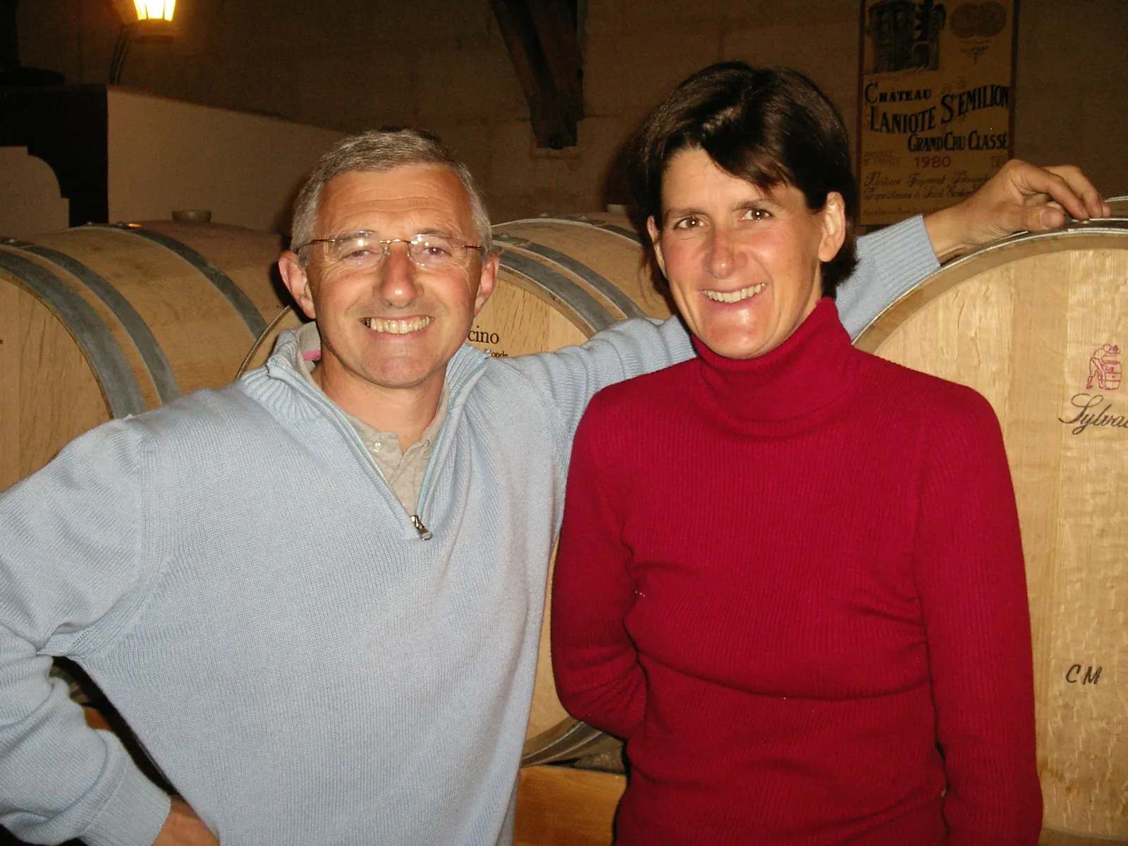 Deux personnes souriantes posant ensemble dans une cave à vin du Château Laniote à Bordeaux, avec des tonneaux de bois en arrière-plan