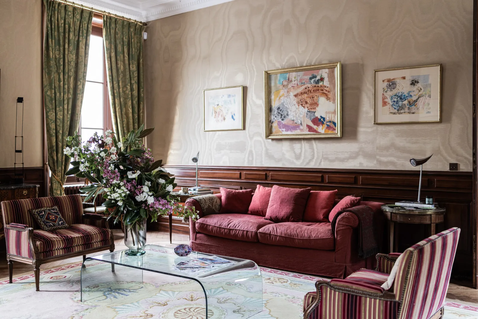 Salon élégant du Château Sénéjac à Bordeaux avec un canapé bordeaux, des fauteuils à rayures, une table basse en verre, des rideaux verts, des œuvres d'art encadrées et des fleurs fraîches.